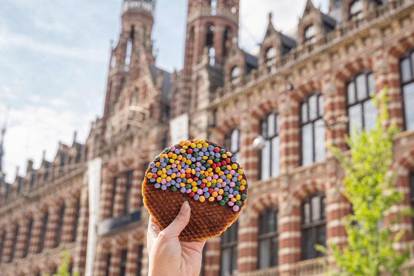 A hand holding a candy-topped stroopwafel in front of a historic Amsterdam building